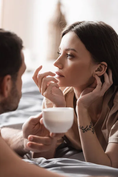 Jeune femme en t-shirt couché sur le lit près de copain flou avec tasse à la maison — Photo de stock