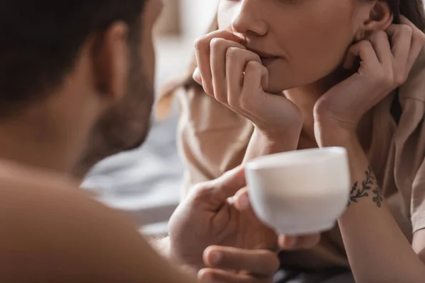 Vista ritagliata della donna tatuata sdraiata sul letto vicino all'uomo sfocato con una tazza di caffè a casa — Foto stock