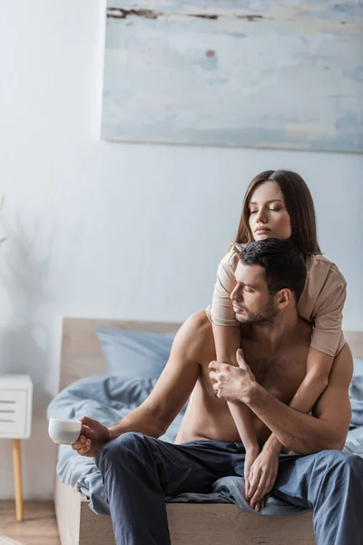 Joven morena cerrando los ojos mientras abraza novio con taza de café en el dormitorio - foto de stock