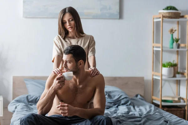 Woman in t-shirt hugging muscular man with coffee cup on bed in morning — Stock Photo