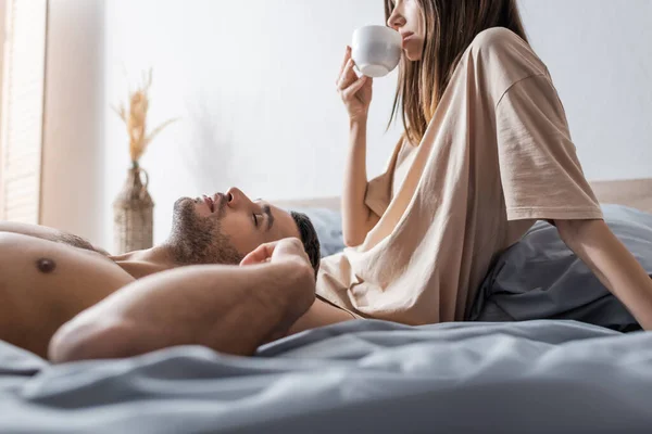 Hombre sexy acostado en la cama cerca de la novia con una taza de café en casa - foto de stock