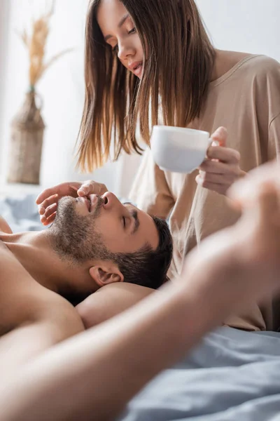 Pretty woman holding blurred cup and touching lips of sexy man on bed — Stock Photo