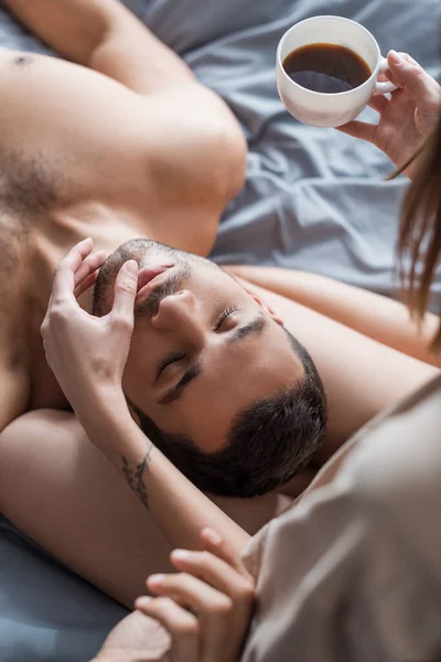 Top view of woman holding cup of coffee and touching lips of shirtless man on bed — Stock Photo