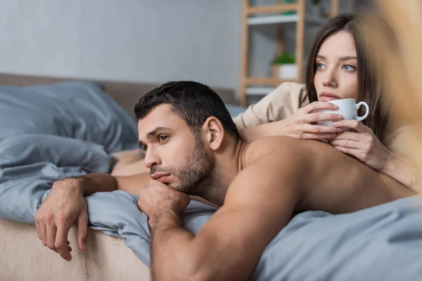 Hombre sin camisa acostado cerca de novia con taza de café en la cama en casa - foto de stock