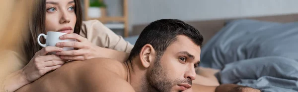 Muskulöser Mann schaut weg, während er neben Freundin mit Tasse auf Bett liegt, Banner — Stockfoto