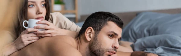 Muscular man looking away while lying near girlfriend with cup on bed, banner — Stock Photo