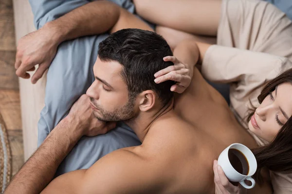 Vista aérea de la mujer sosteniendo café y tocando el pelo de novio muscular en la cama - foto de stock