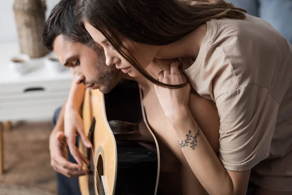 Seitenansicht einer Frau, die ihren hemdlosen Freund umarmt und zu Hause verschwommene Akustikgitarre spielt — Stockfoto