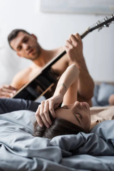 Jovem deitada na cama perto de homem desfocado tocando guitarra acústica — Fotografia de Stock