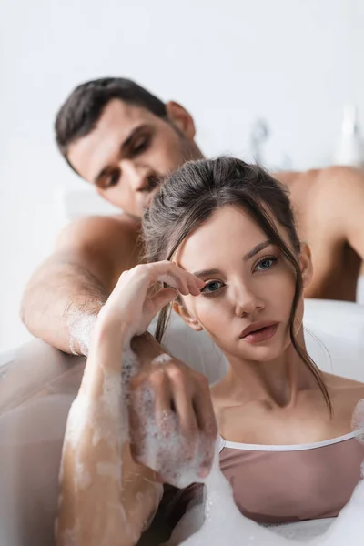 Jovem mulher tomando banho com espuma perto namorado borrado em casa — Fotografia de Stock