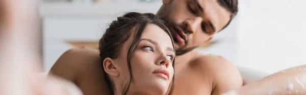 Sexy man looking at brunette girlfriend while taking bath together at home, banner — стоковое фото