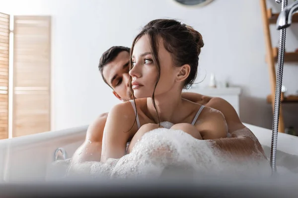 Musculoso hombre en espuma abrazando bonita novia mientras toma baño en casa - foto de stock