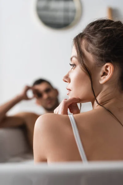 Vista lateral de la mujer joven mirando hacia otro lado mientras toma un baño con novio borroso en casa - foto de stock