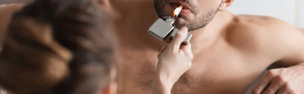 Vista recortada de la mujer sosteniendo encendedor cerca de novio sin camisa con cigarrillo en casa, pancarta - foto de stock