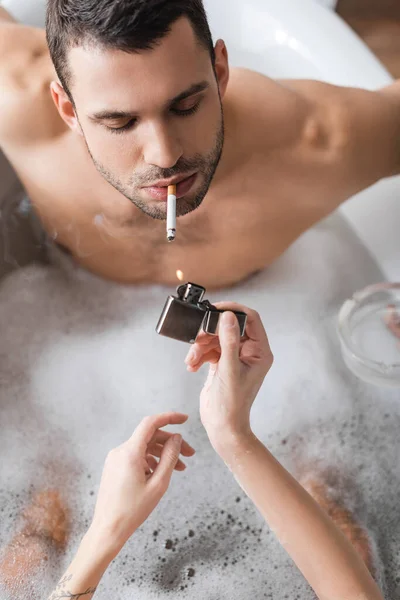 Overhead view of woman holding lighter near muscular boyfriend smoking cigarette in bathtub — Stock Photo