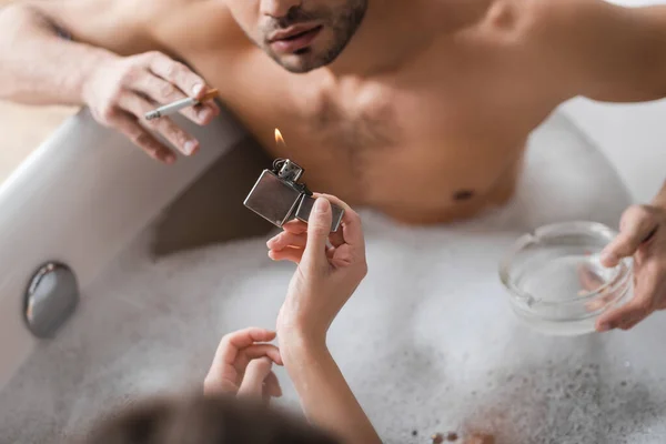 Overhead view of woman holding lighter near blurred boyfriend with cigarette and ashtray in bathtub — Stock Photo