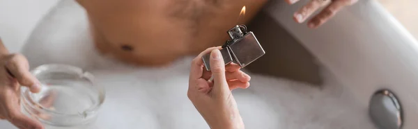 Ausgeschnittene Ansicht einer Frau mit Feuerzeug in der Nähe eines verschwommenen Mannes mit Aschenbecher in Badewanne, Banner — Stockfoto