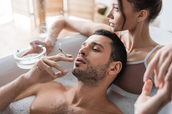 Uomo che tiene la sigaretta vicino alla ragazza con posacenere in bagno con schiuma — Foto stock