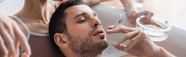 Barded man holding cigarette near girlfriend with ashtray taking bath at home, banner — Stock Photo