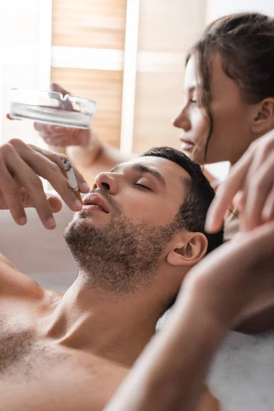 Sexy hombre sosteniendo cigarrillo cerca borrosa novia con cenicero en la bañera - foto de stock
