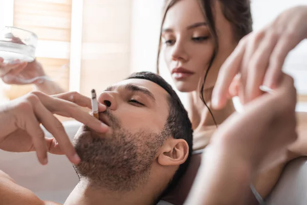 Бородатый мужчина курит сигарету рядом с размытой женщиной, держащей пепельницу в ванной — стоковое фото