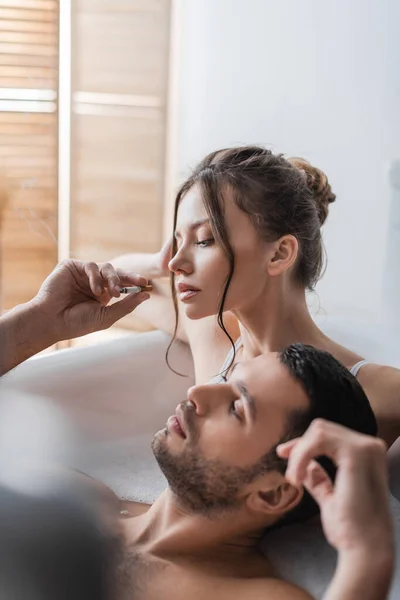 Бородатый мужчина держит сигарету рядом с девушкой в ванной с пеной — стоковое фото