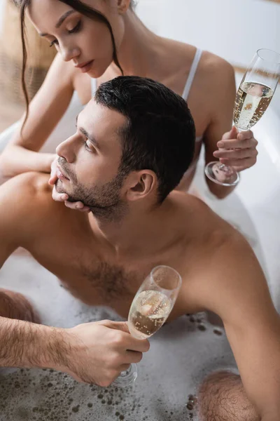 Jeune femme brune touchant son petit ami et tenant du champagne tout en prenant un bain à la maison — Photo de stock