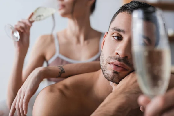 Un hombre sin camisa mirando una copa borrosa de champán cerca de su novia en casa - foto de stock