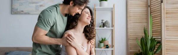 Feliz hombre abrazando alegre y rizado mujer en casa, bandera — Stock Photo