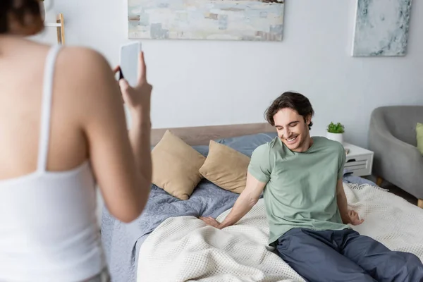 Mujer joven borrosa tomando fotos de novio sonriente en el dormitorio - foto de stock