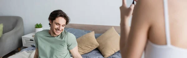 Jeune femme floue prenant une photo de petit ami souriant dans la chambre, bannière — Photo de stock