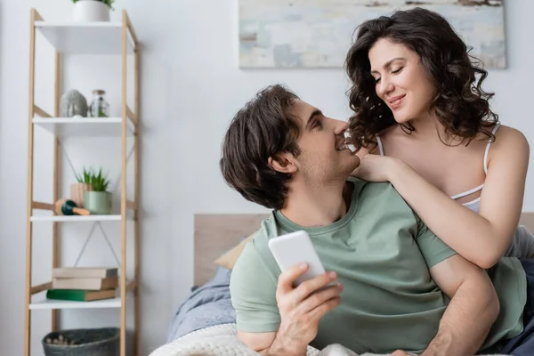 Hombre joven sosteniendo el teléfono inteligente y mirando a la novia en el dormitorio - foto de stock