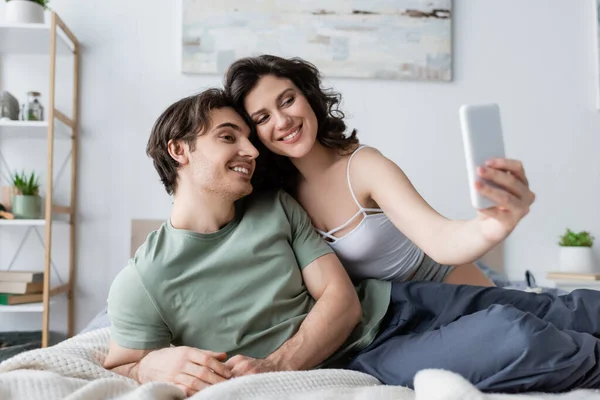 Fröhliches und junges Paar macht Selfie im Schlafzimmer — Stockfoto