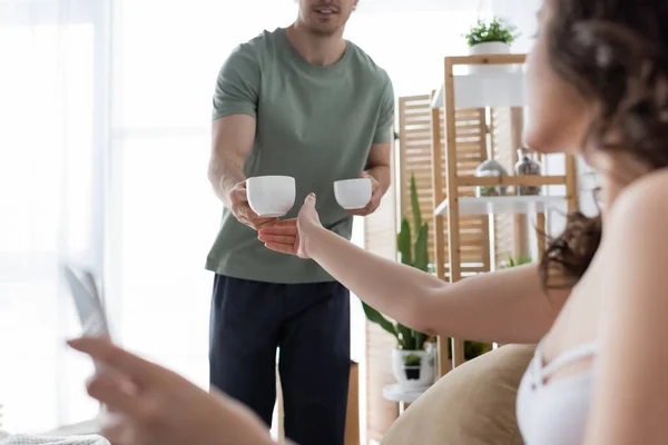 Hombre borroso sosteniendo tazas de café cerca de novia rizada - foto de stock