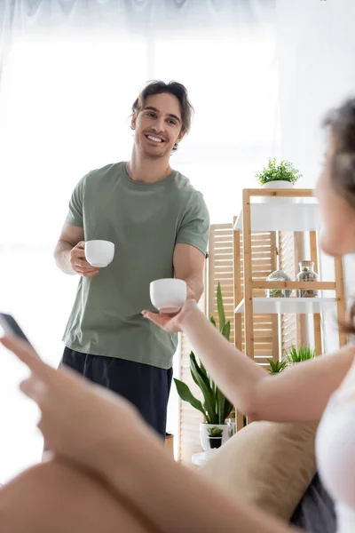 Homme heureux tenant des tasses de café près de petite amie floue — Photo de stock
