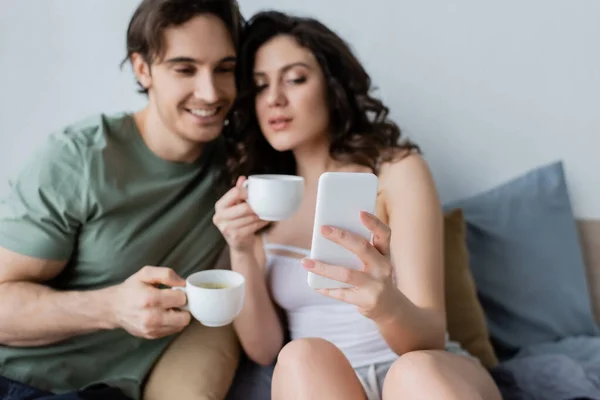 Verschwommenes Paar mit Tassen blickt im Schlafzimmer auf Smartphone — Stockfoto