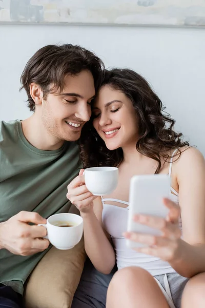Heureux jeune couple avec les yeux fermés tenant des tasses et de prendre selfie — Photo de stock