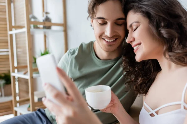 Alegre joven pareja con los ojos cerrados sosteniendo tazas y tomando selfie - foto de stock