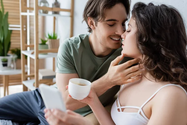 Homem feliz beijando namorada encaracolado com xícara de café e smartphone — Fotografia de Stock