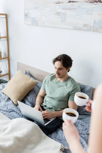 Високий кут зору жінка тримає чашки кави біля позаштатного хлопця, використовуючи ноутбук у ліжку — стокове фото