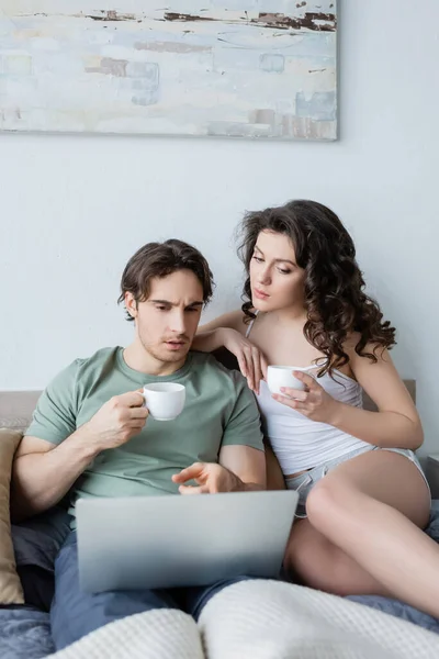 Junge lockige Frau und Mann schauen auf Laptop, während sie Tassen Kaffee im Bett halten — Stockfoto