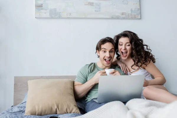 Pareja joven emocionada mirando el ordenador portátil en el dormitorio - foto de stock