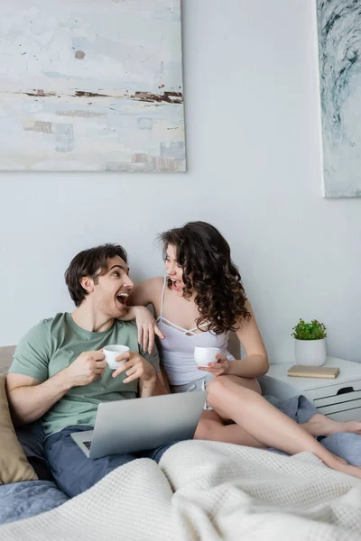 Excitada pareja joven mirándose mientras sostiene tazas cerca de la computadora portátil - foto de stock