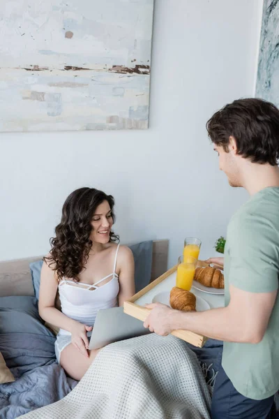 Mann hält Tablett mit leckerem Frühstück neben glücklicher Freundin und Laptop im Bett — Stockfoto