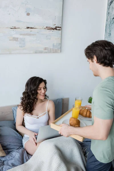 Hombre sosteniendo bandeja con sabroso desayuno cerca de novia feliz y portátil en la cama - foto de stock