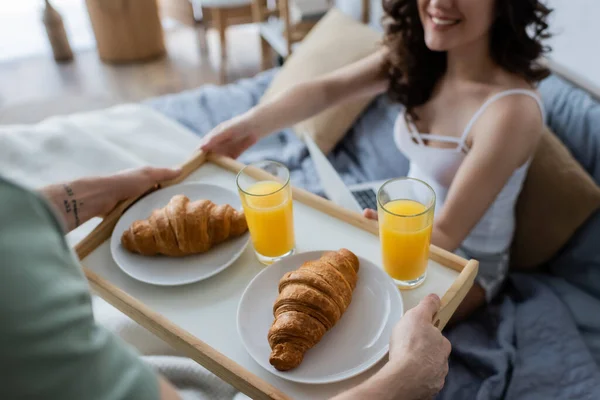 Vista recortada de hombre tatuado sosteniendo bandeja con sabroso desayuno cerca de novia feliz en la cama - foto de stock