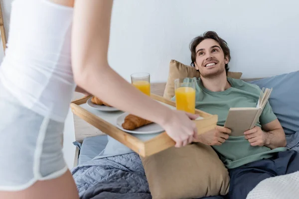 Mujer borrosa sosteniendo bandeja con sabroso desayuno cerca de novio sonriente con libro - foto de stock