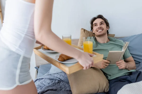 Femme floue tenant plateau avec petit déjeuner savoureux près copain souriant avec livre — Photo de stock