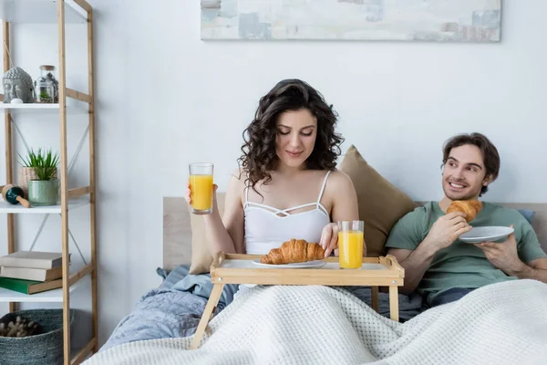 Mulher feliz segurando vidro de suco de laranja perto de homem alegre com croissant — Fotografia de Stock