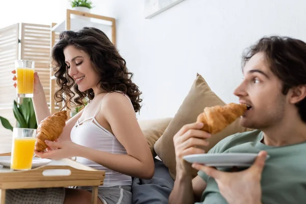Blurred man eating croissant and looking at happy woman during breakfast in bed — Stock Photo